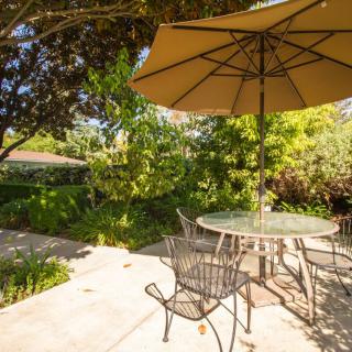 A sunny patio with a table, chairs, and an umbrella surrounded by lush greenery.