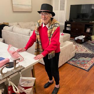 An elderly individual showcasing a stylish outfit with a red sweater and plaid scarf, standing in a cozy living room.