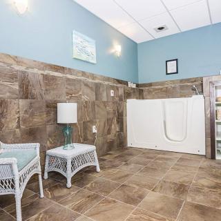 A serene waiting area featuring a walk-in bathtub, wicker chairs, and a lamp against a backdrop of tile flooring and muted wall colors.