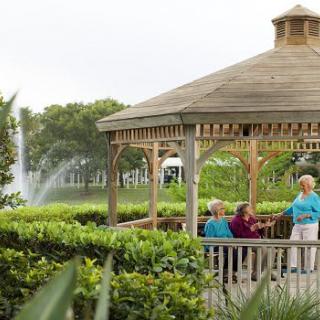 A group of people enjoying a conversation under a wooden gazebo in a lush garden setting with a fountain in the background.