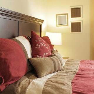 A cozy bedroom with a neatly arranged bed, featuring multiple pillows and a warm lamp in the background.