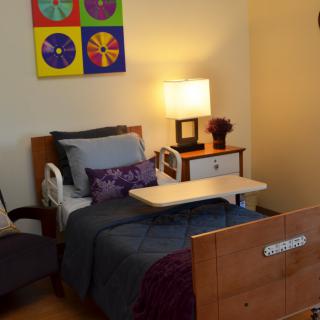 A cozy bedroom with a hospital bed, colorful artwork, and a bedside table with a lamp.
