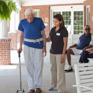 An elderly man using a cane is assisted by a caregiver in an outdoor setting at a nursing home.