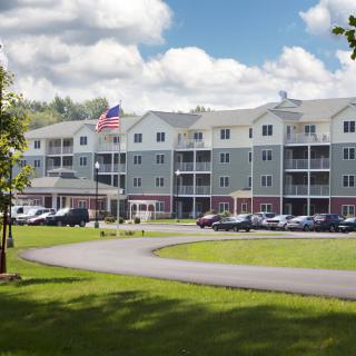 A multi-story residential building with a well-maintained lawn and parking lot under a cloudy sky.