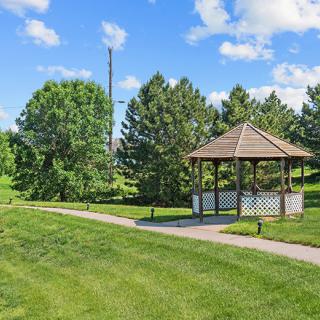 A peaceful park scene with a wooden gazebo, surrounded by lush green grass and trees under a bright blue sky.