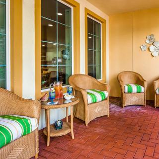 A cozy patio with wicker chairs featuring green and white striped cushions, a small table with drinks, and large windows.