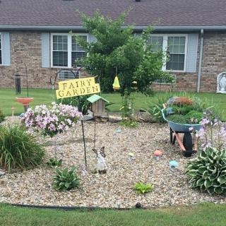 A charming garden with a variety of plants and flowers, featuring a sign and wheelbarrow in front of a house.