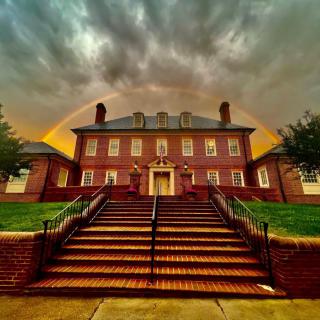A majestic brick building framed by a vibrant rainbow and set against a dramatic cloudy sky.