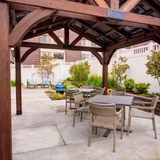 A cozy outdoor patio area with tables and chairs under a wooden gazebo, surrounded by a well-maintained garden.
