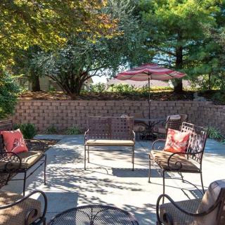 A cozy outdoor patio area with metal furniture and a garden umbrella, surrounded by trees and a brick wall.