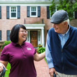 A caregiver assists an elderly man with a walking aid outside a house.