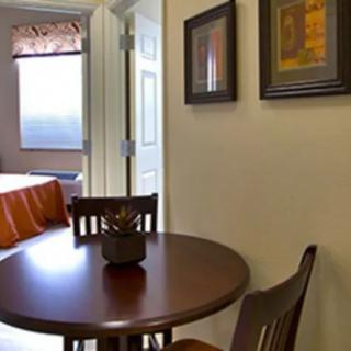 A cozy room interior featuring a small round dining table with chairs, artwork on the walls, and a glimpse of a bed near a window.