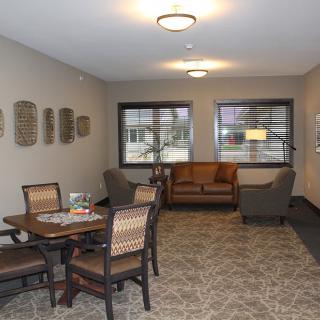A cozy living room with a seating area, including a sofa, chairs, and a table. The space is well-lit with natural light coming through the windows.