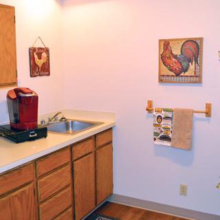 A small kitchen area featuring a coffee maker and wood cabinets, adorned with wall art and a towel rack.