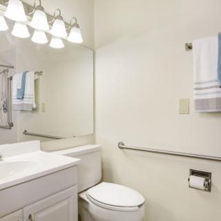 A clean and bright bathroom with a toilet, sink, and mirror, featuring a towel rack and overhead lighting.
