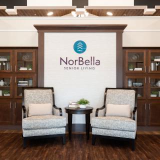A welcoming lobby area with two upholstered chairs and a table, featuring the NorBella logo on the wall.