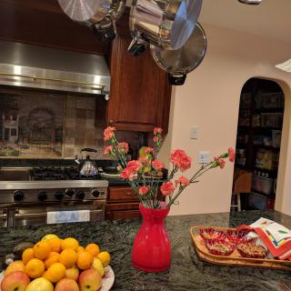 A kitchen with a granite countertop displaying a bowl of oranges, a red vase with flowers, and snacks in a basket.
