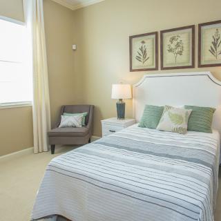 A cozy bedroom featuring a neatly made bed with striped bedding, decorative pillows, and three framed artworks above the headboard. A comfortable chair sits in the corner near a window, with a lamp on the bedside table completing the room's warm ambiance.