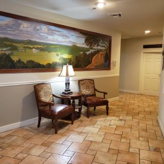 A hallway with tiled flooring, featuring a large landscape painting, two chairs, and a table with a lamp.