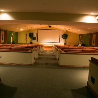 An empty auditorium with rows of wooden seats facing a large projector screen.