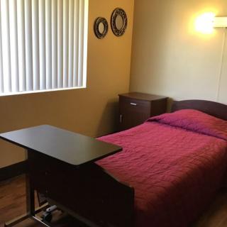 A minimalist bedroom with a single bed featuring a red bedspread and an overbed table, accompanied by wall decorations and window blinds.