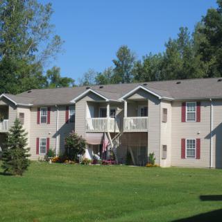 A two-story apartment building surrounded by trees and green lawn on a clear day.