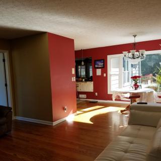 A cozy living room with red walls and hardwood flooring, featuring a dining area and chandelier bathed in natural sunlight.
