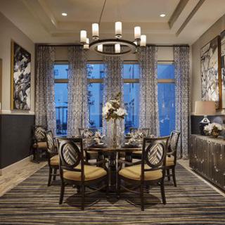 A beautifully decorated dining room featuring a round table surrounded by chairs, elegant chandelier, artwork on the walls, and large windows with patterned curtains.