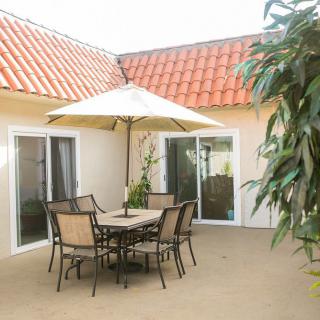 A cozy patio area with a table, chairs, and an umbrella, surrounded by plants and enclosed by sliding glass doors under a terracotta roof.