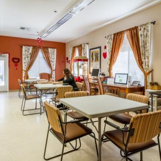A cozy dining area with tables and chairs, decorated with curtains and wall hangings, featuring a computer station by the window.