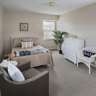 A cozy bedroom featuring a neatly made bed, a white dresser, and a comfortable chair, complemented by soft lighting and minimalist decor.