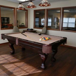 A well-lit game room featuring a classic pool table with a cue stick and racked balls on a wooden floor.