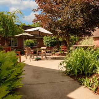 A serene outdoor garden area with patio furniture shaded by an umbrella, surrounded by lush greenery.