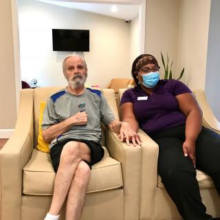 An elderly man and a caregiver sitting together on a sofa; the caregiver is wearing a mask.
