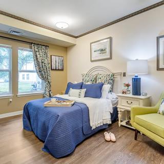 A cozy bedroom with a blue-themed bedspread, decorative pillows, and a green armchair next to a window with curtains.