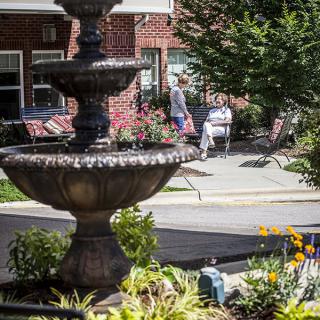 A tranquil outdoor setting featuring a tiered fountain, surrounded by lush landscaping and vibrant flowers, with benches in the background.