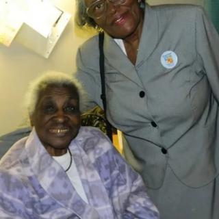 Two smiling individuals posing together indoors, with one seated and the other standing.