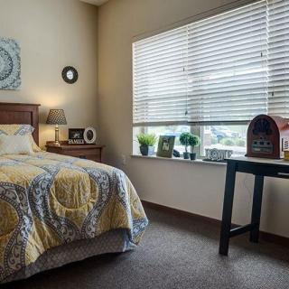 A cozy bedroom with a neatly made bed, a side table with decor items, and a window allowing natural light into the room.