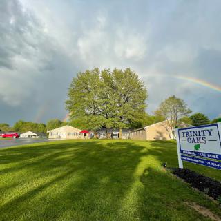 A landscape view of a building with a rainbow in the cloudy sky, a large tree, and a sign on the green lawn.