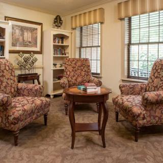 A cozy living room with patterned armchairs and a small wooden table, flanked by windows and a bookshelf.