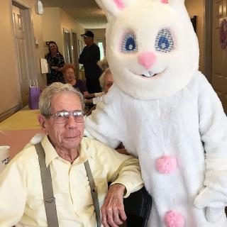 An elderly man sitting next to a person in an Easter Bunny costume at an indoor event.