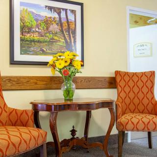 A cozy corner with two patterned orange chairs, a wooden table with a vase of yellow flowers, and a landscape painting on the wall.