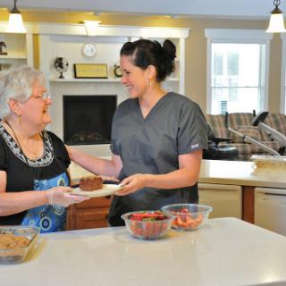 A caregiver serving food to an elderly woman in a warm kitchen setting.
