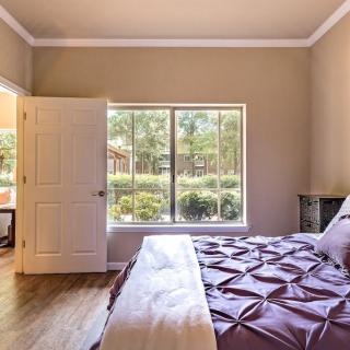 A cozy bedroom with a large window overlooking a garden, featuring a bed with purple bedding and an open door leading to another room.