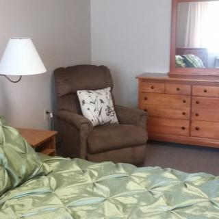 A cozy bedroom featuring a green bedspread, an armchair with a decorative pillow, a wooden dresser with a mirror, and a lamp.