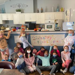 A group of children wearing masks gather indoors for a celebration in front of a decorated banner in a kitchen setting.