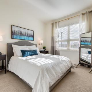 A modern bedroom with a neatly made bed, artwork above the headboard, and a large window allowing natural light to fill the room.