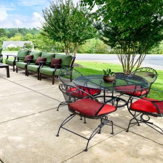 A charming outdoor patio setup featuring a round table with red-cushioned chairs and a line of sofas with green cushions, set against a lush backdrop of trees and grass.