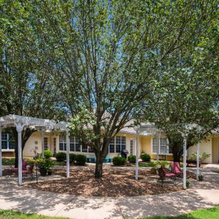 A scenic outdoor area featuring a garden with trees, chairs, and a pergola in front of a building.