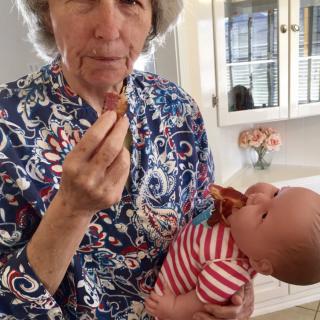 A woman holds a baby doll and humorously pretends to feed it bacon in a kitchen.